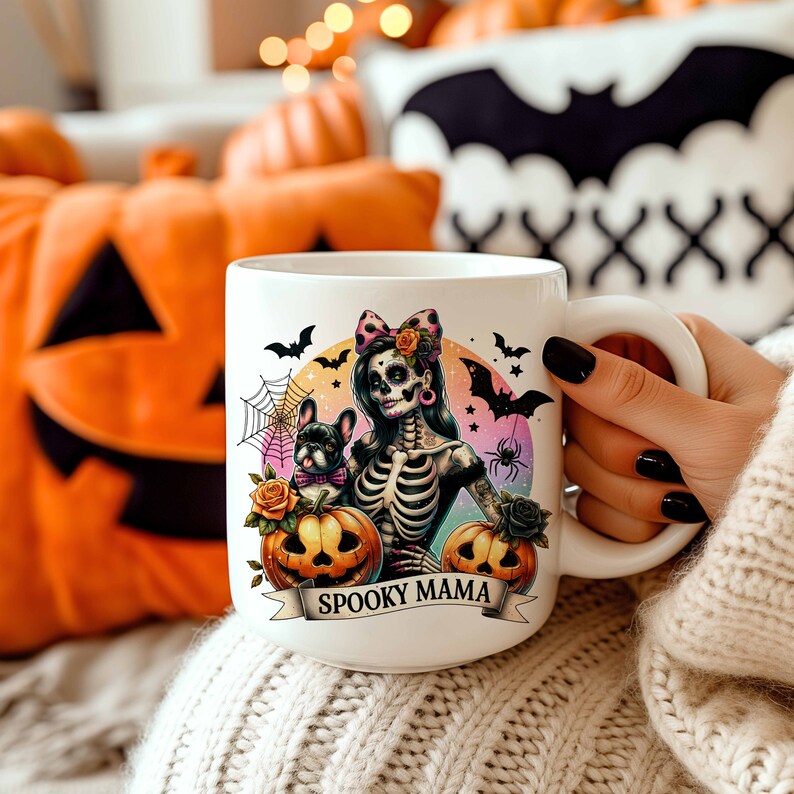 Peut inclure: Une tasse en c&eacute;ramique blanche avec une illustration color&eacute;e d'une femme squelette au visage de cr&acirc;ne, portant un n&oelig;ud rose et tenant un petit chien noir et blanc. La tasse porte le texte "Spooky Mama" en bas.
