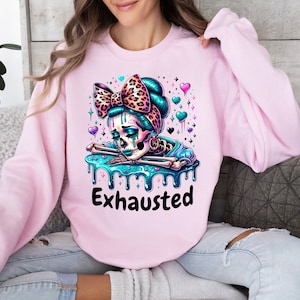 May include: A light pink sweatshirt featuring a colorful graphic of a skeleton with a leopard print bow and the word "Exhausted" in black. The skeleton has teal hair and is surrounded by hearts and stars.
