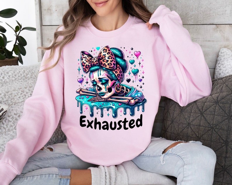 May include: A light pink sweatshirt featuring a colorful graphic of a skeleton with a leopard print bow and the word "Exhausted" in black. The skeleton has teal hair and is surrounded by hearts and stars.
