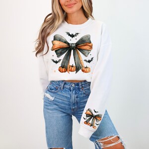 May include: White sweatshirt with a black and orange bow, bats, and pumpkins. The bow has a black and white polka dot pattern.