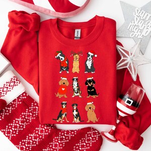 May include: A red crewneck sweatshirt featuring nine cartoon dogs in festive attire, including Santa hats and reindeer antlers. The dogs are arranged in three rows. The sweatshirt is surrounded by Christmas decorations, including a stocking and a wine glass.