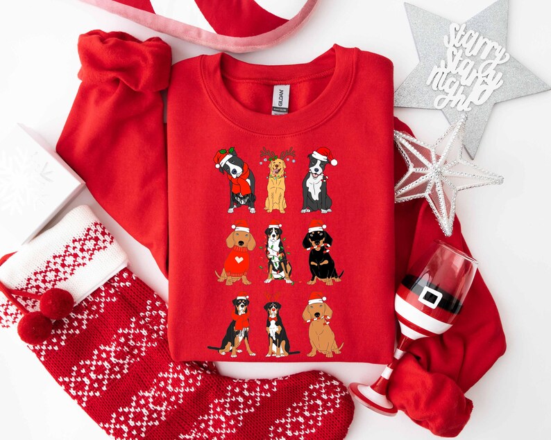 May include: A red crewneck sweatshirt featuring nine cartoon dogs in festive attire, including Santa hats and reindeer antlers. The dogs are arranged in three rows. The sweatshirt is surrounded by Christmas decorations, including a stocking and a wine glass.
