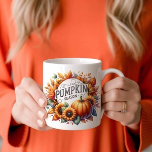 Pu&ograve; includere: Tazza in ceramica bianca con un'illustrazione colorata di zucche, girasoli e foglie autunnali. La tazza ha il testo "Pumpkin Season" in un font corsivo nero.