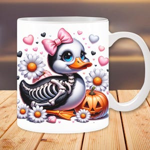 May include: White ceramic mug with a cartoon image of a duck with a pink bow, a skeleton, and a jack-o-lantern. The duck is surrounded by hearts and flowers.