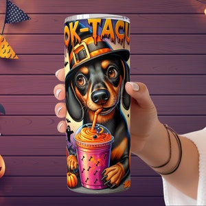 Może przedstawiać: Czarny i brązowy szczeniak rasy dachshund w czarnym kapeluszu czarownicy trzymający r&oacute;żowy i pomarańczowy nap&oacute;j na Halloween. Na kubku widnieje napis "Boo-Tacular".