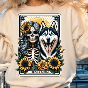 Puede incluir: Una sudadera blanca con un dise&ntilde;o gr&aacute;fico de una mujer esqueleto con girasoles y un perro husky. El texto "HUSKY MOM" est&aacute; impreso debajo de la imagen.