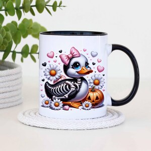May include: White ceramic coffee mug with a black rim and handle. The mug features a cartoon illustration of a duck with a pink bow, a skeleton, and a jack-o-lantern. The background is decorated with hearts and flowers.