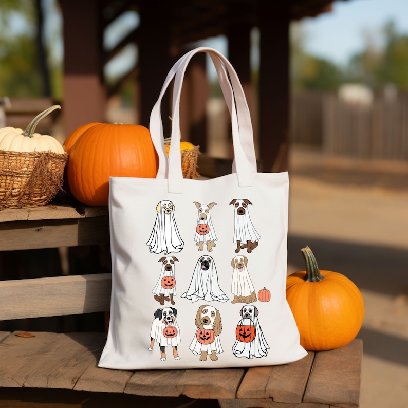 Pu&ograve; includere: Una borsa tote bianca con un design a tema Halloween di cani vestiti da fantasmi, ognuno con una lanterna a forma di zucca. La borsa ha due manici bianchi ed &egrave; esposta con zucche su una superficie di legno.