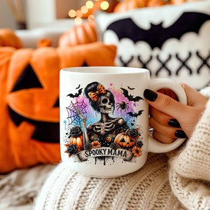 May include: White ceramic mug with a colorful illustration of a skeleton woman with a bow in her hair, holding a pumpkin. The illustration is surrounded by bats, a spider web, and the text "Spooky Mama".