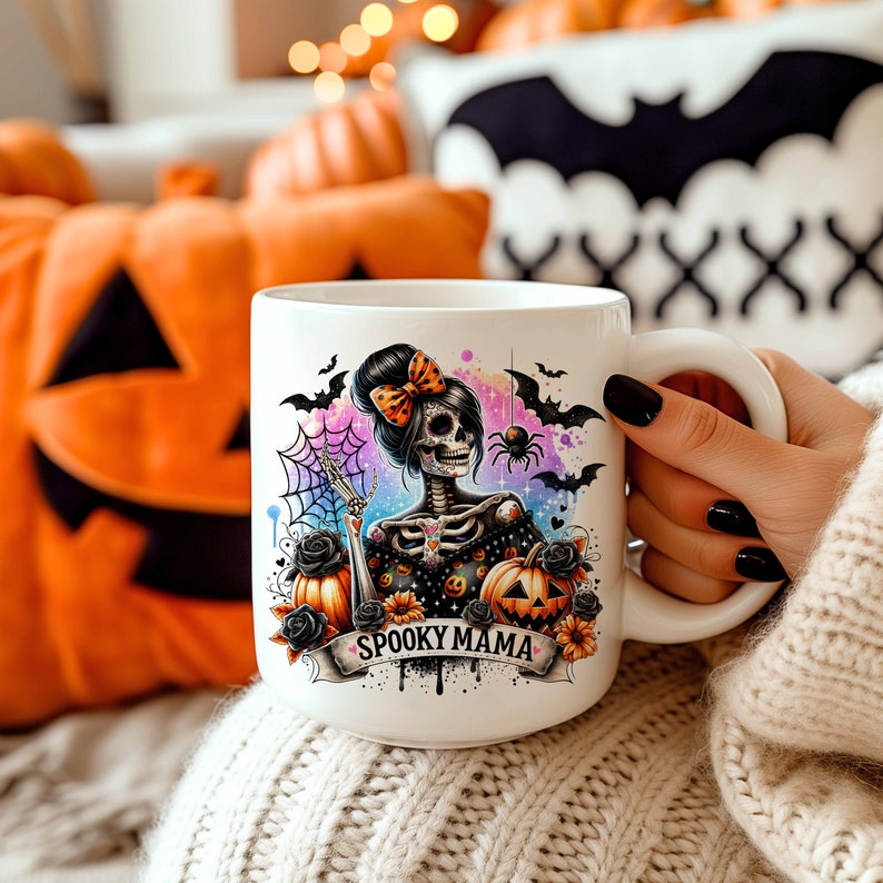 May include: White ceramic mug with a colorful illustration of a skeleton woman with a bow in her hair, holding a pumpkin. The illustration is surrounded by bats, a spider web, and the text "Spooky Mama".