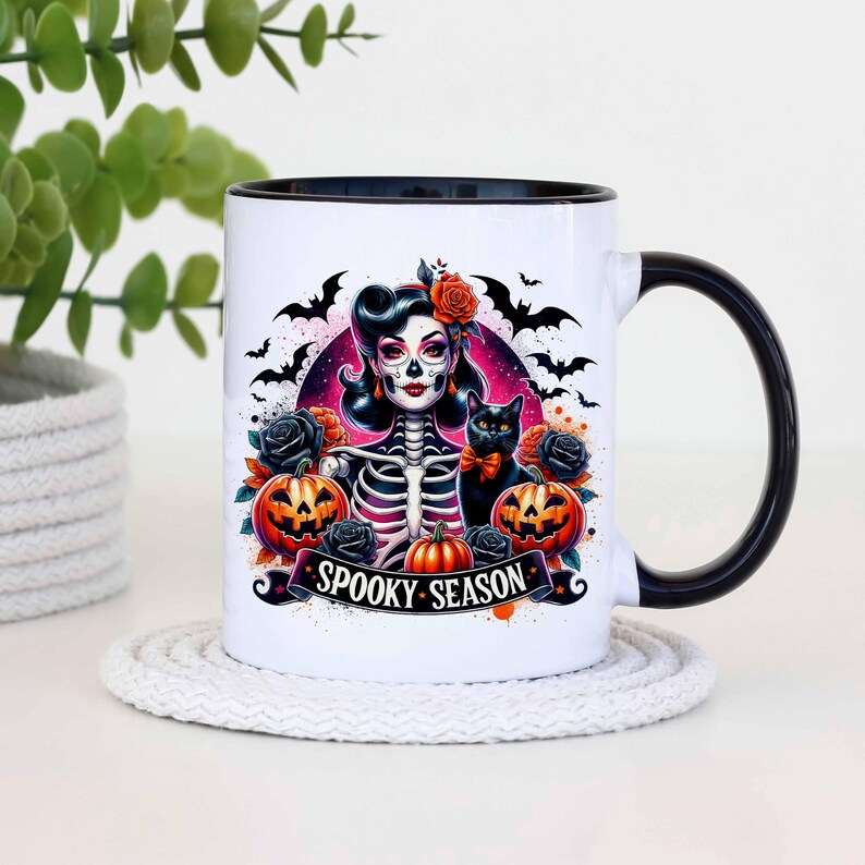 May include: White ceramic mug with a black rim and handle. The mug features a colorful illustration of a skeleton woman with a black cat, pumpkins, and bats. The text "Spooky Season" is printed on the mug.