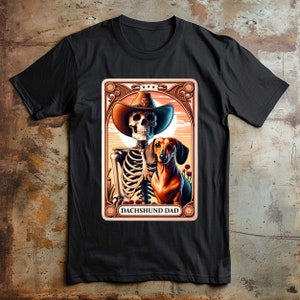 Op de afbeelding: Een zwart T-shirt met een afbeelding van een skelet dat een cowboyhoed draagt en een teckel vasthoudt. De afbeelding is in vintage stijl met een gouden frame en de tekst "Dachshund Dad".