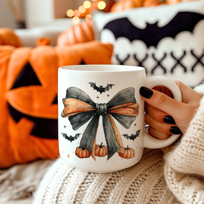 May include: A white ceramic mug with a black and orange bow and bats printed on it. The mug is sitting on a white knitted blanket.