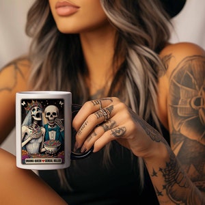 May include: White ceramic mug with a black handle. The mug features a colorful illustration of two skeletons dressed as a bride and groom. The text "Drama Queen &hearts; Cereal Killer" is printed on the mug.