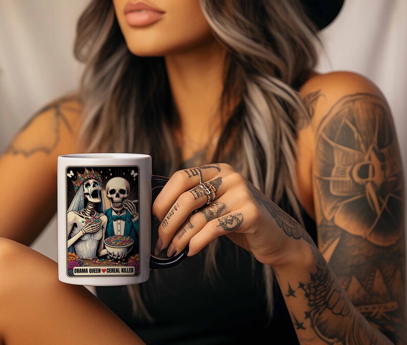 May include: White ceramic mug with a black handle. The mug features a colorful illustration of two skeletons dressed as a bride and groom. The text "Drama Queen &hearts; Cereal Killer" is printed on the mug.
