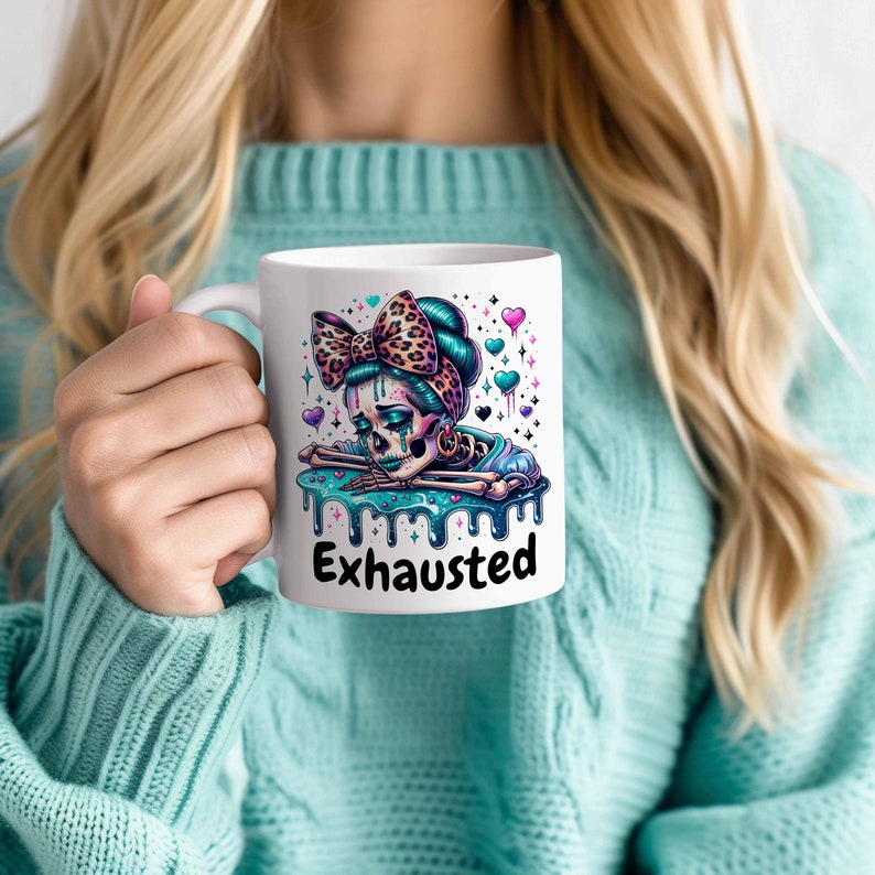 May include: White ceramic mug featuring a colorful illustration of a skeleton with a bow and the word "Exhausted". The design includes teal, pink, and purple accents. The mug is held by a person wearing a teal sweater.