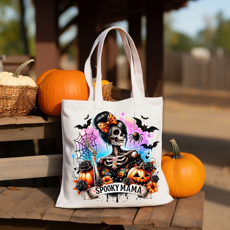 May include: White tote bag with a colorful illustration of a skeleton woman with a bow in her hair. The illustration is surrounded by bats, spiders, and pumpkins. The text "Spooky Mama" is printed on the bag.