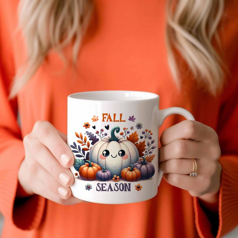 May include: White ceramic mug featuring a cute cartoon pumpkin design with the words "FALL SEASON". The design includes pumpkins in various sizes and colors, surrounded by fall foliage. The mug is held by a person wearing an orange sweater.