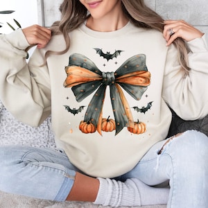 May include: A cream-colored sweatshirt with a black and orange bow with a watercolor effect. The bow is decorated with small black bats and orange pumpkins.