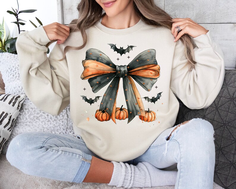 May include: A cream-colored sweatshirt with a black and orange bow with a watercolor effect. The bow is decorated with small black bats and orange pumpkins.