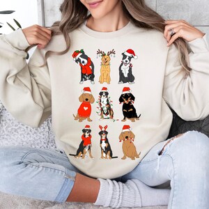 May include: A cream-colored sweatshirt featuring a festive design of various dog breeds dressed in Christmas attire. The dogs are wearing Santa hats, scarves, and reindeer antlers. The graphic is a fun, seasonal design.