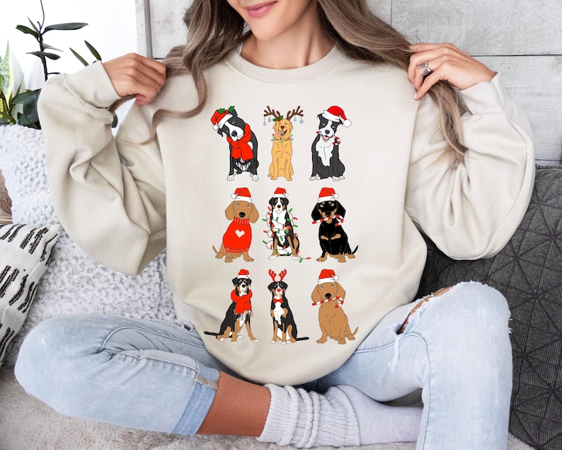 May include: A cream-colored sweatshirt featuring a festive design of various dog breeds dressed in Christmas attire. The dogs are wearing Santa hats, scarves, and reindeer antlers. The graphic is a fun, seasonal design.