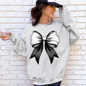 Puede incluir: Una sudadera gris con un gran lazo negro y blanco en la parte delantera.