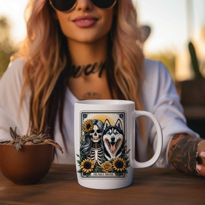 Puede incluir: Taza de cer&aacute;mica blanca con una ilustraci&oacute;n colorida de una mujer esqueleto con una corona de girasol y un perro husky. El texto "HUSKY MOM" est&aacute; impreso en la parte inferior de la taza.
