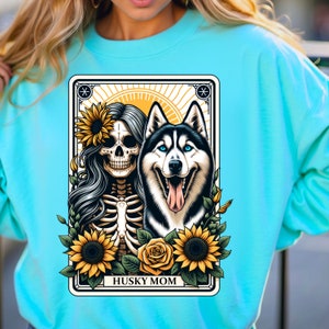 Puede incluir: Una sudadera turquesa con un dise&ntilde;o gr&aacute;fico de un esqueleto con girasoles y un perro husky. El gr&aacute;fico est&aacute; rodeado por un marco negro con el texto "Husky Mom".