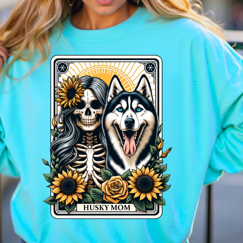 Puede incluir: Una sudadera turquesa con un dise&ntilde;o gr&aacute;fico de un esqueleto con girasoles y un perro husky. El gr&aacute;fico est&aacute; rodeado por un marco negro con el texto "Husky Mom".