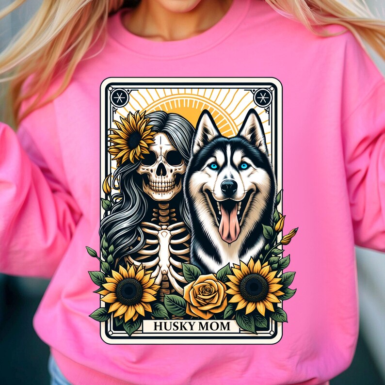 Puede incluir: Una sudadera rosa con un dise&ntilde;o gr&aacute;fico de una mujer esqueleto con girasoles y un perro husky. El gr&aacute;fico est&aacute; enmarcado en una tarjeta de estilo vintage con el texto "Husky Mom".