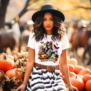 Peut inclure: Un t-shirt blanc avec un graphique color&eacute; d'une femme squelette tenant des citrouilles et un petit chien. Le texte "Spooky Mama" est imprim&eacute; sur une banni&egrave;re sous le graphique.
