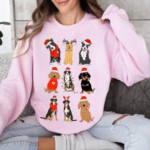 May include: A light pink sweatshirt featuring nine cartoon dogs in festive holiday attire. Each dog wears a Santa hat, and some have scarves or antlers. The dogs are various breeds and colors, creating a cheerful, seasonal design.