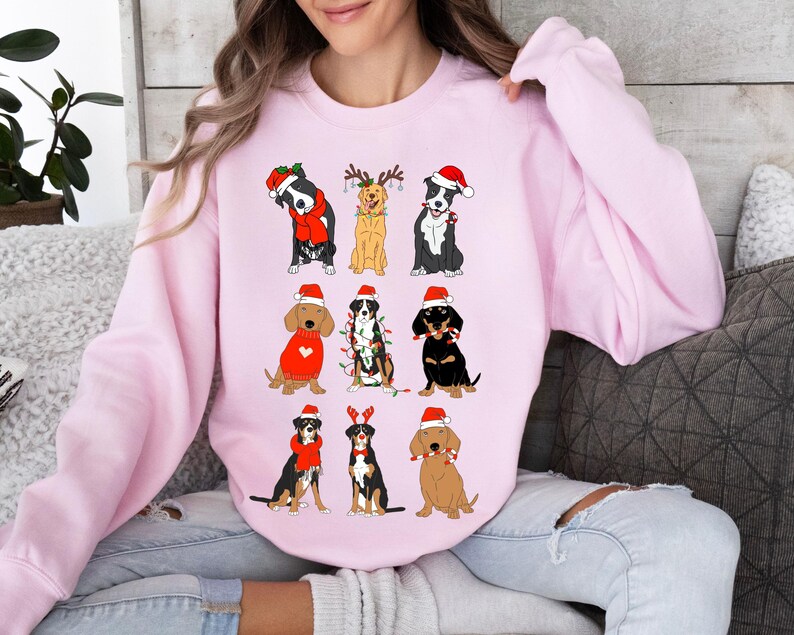 May include: A light pink sweatshirt featuring nine cartoon dogs in festive holiday attire. Each dog wears a Santa hat, and some have scarves or antlers. The dogs are various breeds and colors, creating a cheerful, seasonal design.