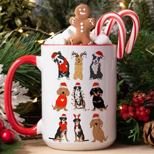 May include: A white ceramic mug with a red handle and rim, decorated with cartoon dogs in Christmas attire. The mug is filled with marshmallows, a gingerbread man, and candy canes. Festive holiday theme.