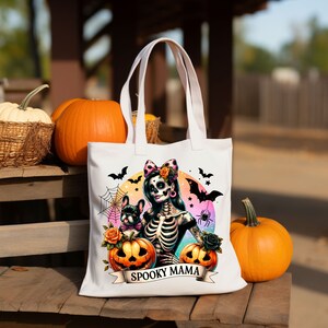 Peut inclure: Sac fourre-tout blanc avec une illustration color&eacute;e d'une femme squelette avec un n&oelig;ud rose dans les cheveux et un chien. La femme tient une citrouille et le texte "Spooky Mama" est imprim&eacute; sur le sac.