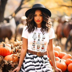 Pu&ograve; includere: Una maglietta bianca con un design a tema Halloween con illustrazioni di cani vestiti da fantasmi e con zucche. La modella indossa un cappello nero, una gonna a righe e una cintura marrone. Le zucche sono in primo piano.