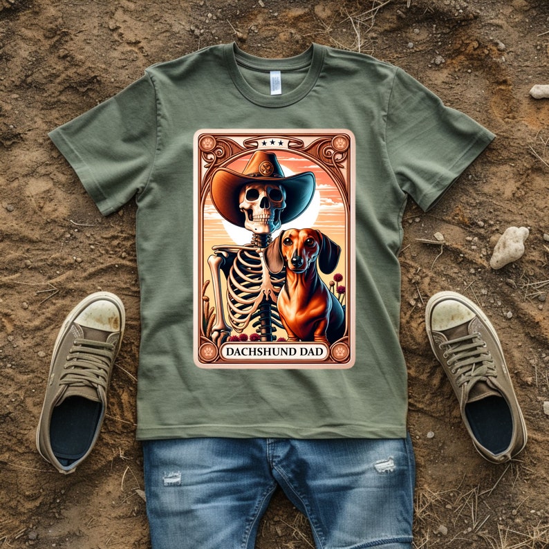 Op de afbeelding: Een groen T-shirt met een afbeelding van een skelet dat een cowboyhoed draagt en een teckel vasthoudt. De afbeelding is omgeven door een decoratief kader en de tekst "Dachshund Dad" staat onderaan.