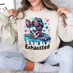 May include: Beige sweatshirt featuring a colorful graphic of a skeleton with a teal bow, teal hair, and a teal liquid design. The word "Exhausted" is printed in black below the graphic. The design includes hearts and stars.