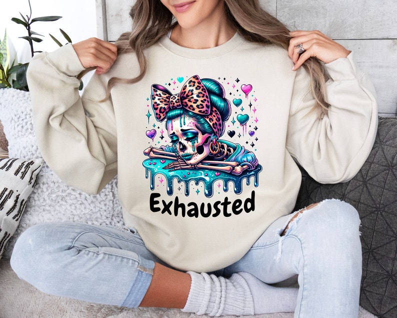 May include: Beige sweatshirt featuring a colorful graphic of a skeleton with a teal bow, teal hair, and a teal liquid design. The word "Exhausted" is printed in black below the graphic. The design includes hearts and stars.