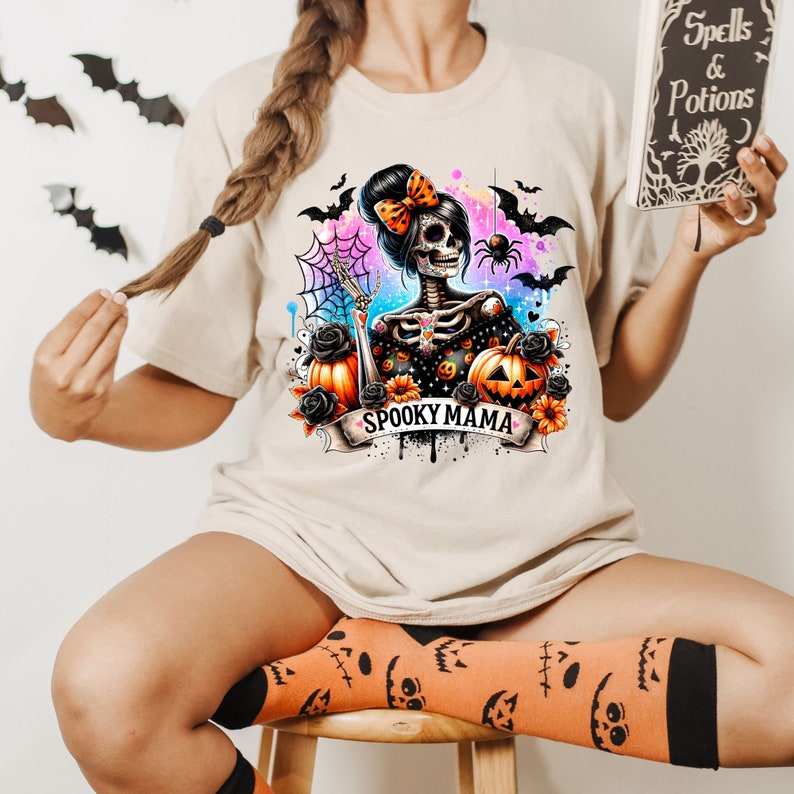 May include: A beige t-shirt with a colorful graphic of a skeleton woman with a bow in her hair, holding a pumpkin and surrounded by bats, spiders, and flowers. The text "Spooky Mama" is written on a banner below the graphic.