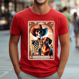 Op de afbeelding: Rood T-shirt met een afbeelding van een skelet dat een cowboyhoed draagt en een teckel vasthoudt. De afbeelding is in een vintage stijl frame met de tekst "Dachshund Dad".