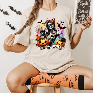 Peut inclure: Un t-shirt blanc avec une illustration color&eacute;e d'une femme squelette tenant une citrouille et un petit chien. Le texte "Spooky Mama" est imprim&eacute; sur une banni&egrave;re sous l'illustration.