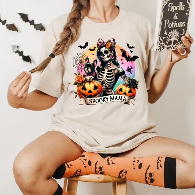 Peut inclure: Un t-shirt blanc avec une illustration color&eacute;e d'une femme squelette tenant une citrouille et un petit chien. Le texte "Spooky Mama" est imprim&eacute; sur une banni&egrave;re sous l'illustration.
