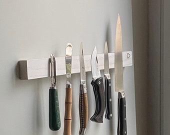 Peut inclure: Un porte-couteaux magnétique blanc avec six couteaux attachés. Les couteaux ont des manches en bois et noirs. Le porte-couteaux a une forme de cœur sculptée dans le bois.
