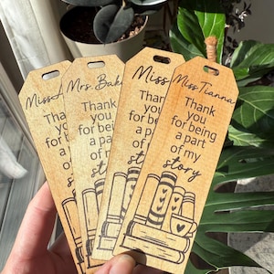 May include: Wooden bookmarks with a natural finish. Each bookmark features a unique design with the text "Thank you for being a part of my story" and a stack of books with a coffee cup. The bookmarks are personalized with names.