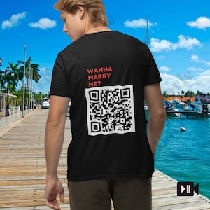 Puede incluir: Una camiseta negra con el texto "Wanna Marry Me?" y un código QR impreso en la espalda. La persona que lleva la camiseta está de pie en un muelle de madera con vistas a un puerto deportivo.