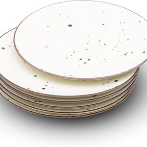 May include: A stack of six off-white ceramic plates with a speckled design and a brown rim. The plates are round and have a simple, elegant design. The plates are stacked on top of each other.