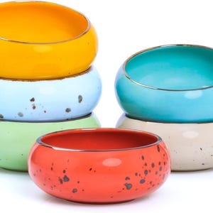May include: A collection of colorful ceramic bowls in various sizes. The bowls are stacked and arranged, showcasing a palette of yellow, light blue, green, red, and beige. Each bowl has a metallic gold rim and speckled detailing.