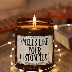 May include: A lit brown glass jar candle with the text "SMELLS LIKE YOUR CUSTOM TEXT" on a white label. The candle sits on a wooden coaster, surrounded by string lights. The candle's label also includes the text "Gifts Made of Love and Humor".
